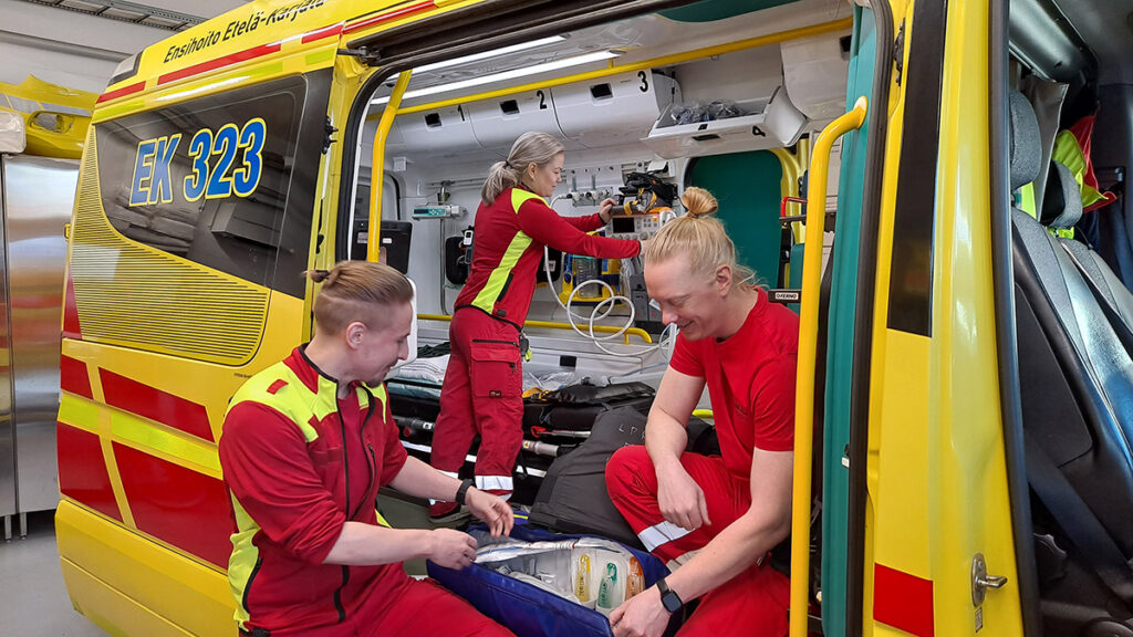 Ambulanssin avoimen oven edessä kyykistyneenä kaksi ensihoitajaa tutkimassa varustelaukun sisältöä ja yksi ensihoitaja järjestelee varusteita ambulanssin sisällä. 