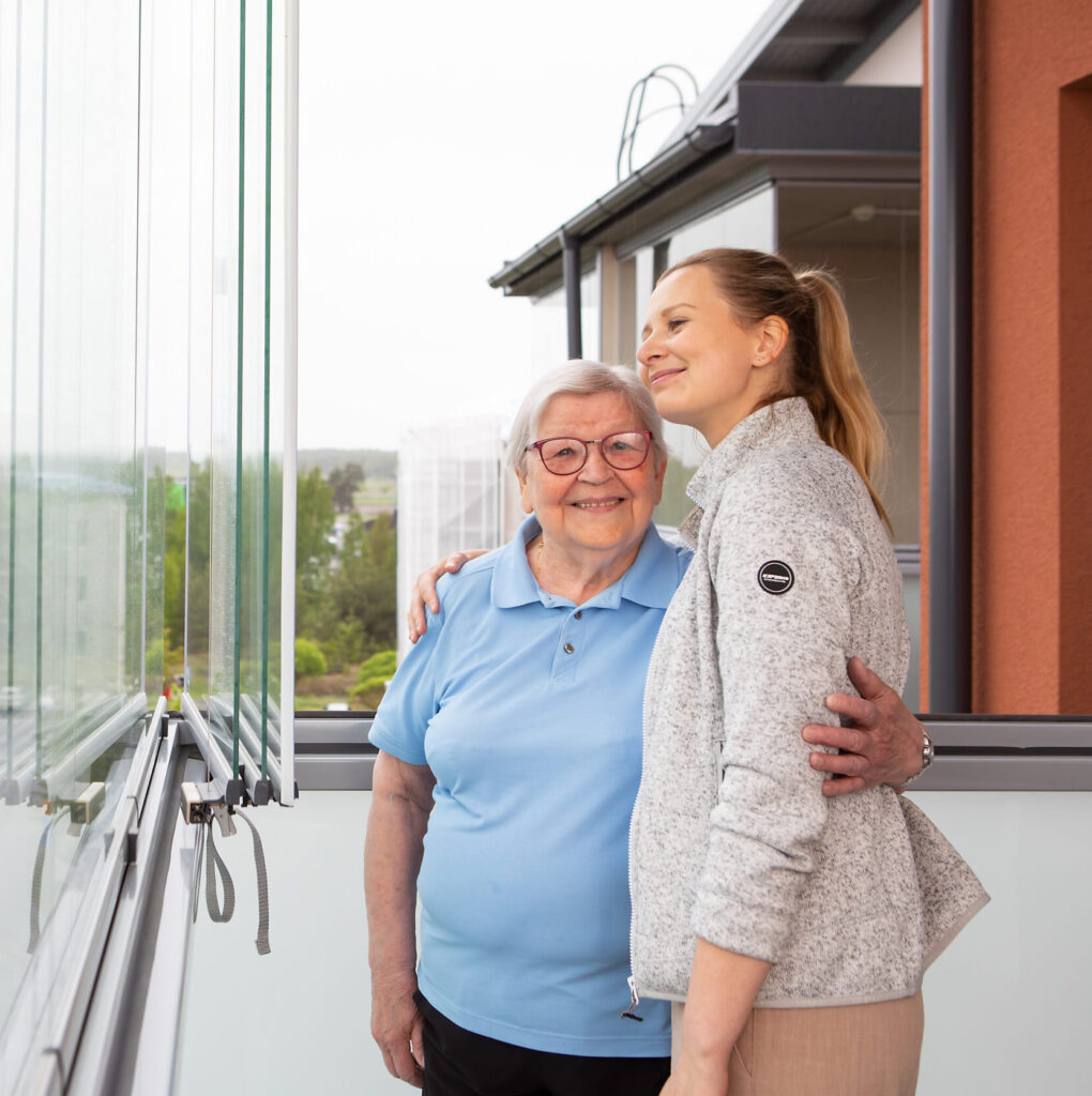 Hymyilevät naiset, iäkäs asiakas ja hoitaja seisovat kerrostalon parvekkeella toistensa kainalossa. Iäkäs nainen katsoo kameraan.